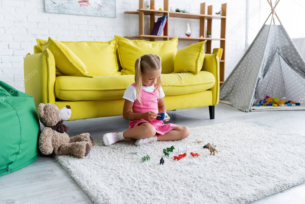 preschooler girl sitting on carpet and playing with toy in modern playroom 
