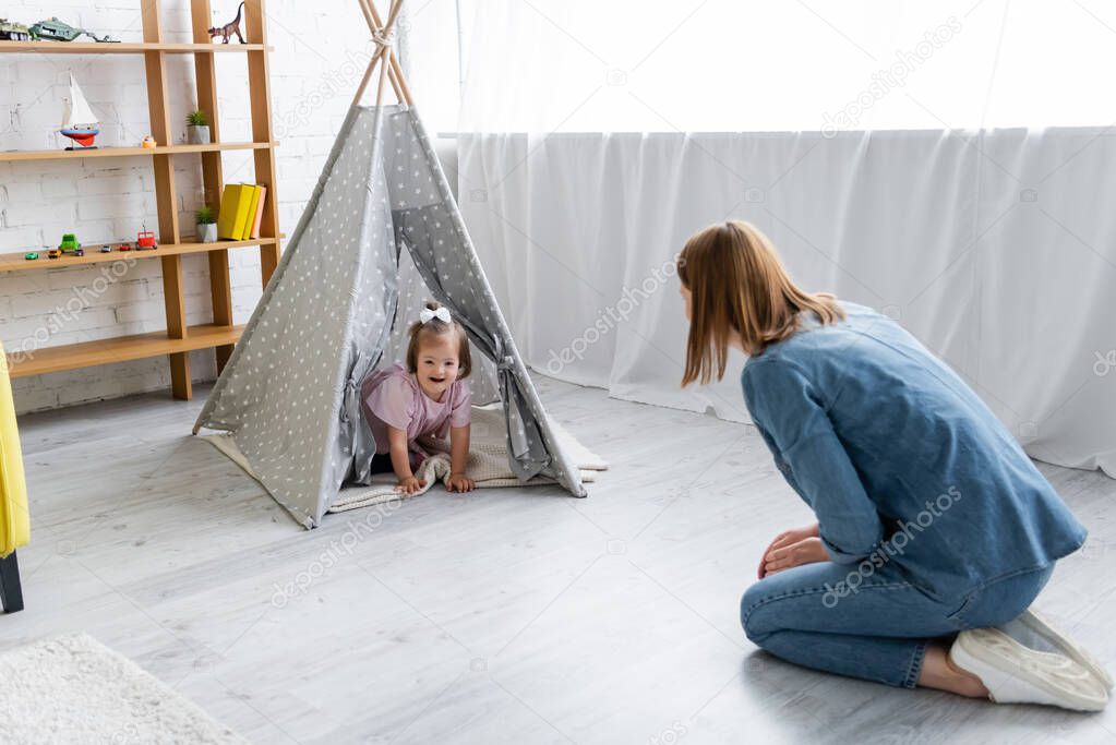 kindergarten teacher looking at happy kid with down syndrome sitting in tipi