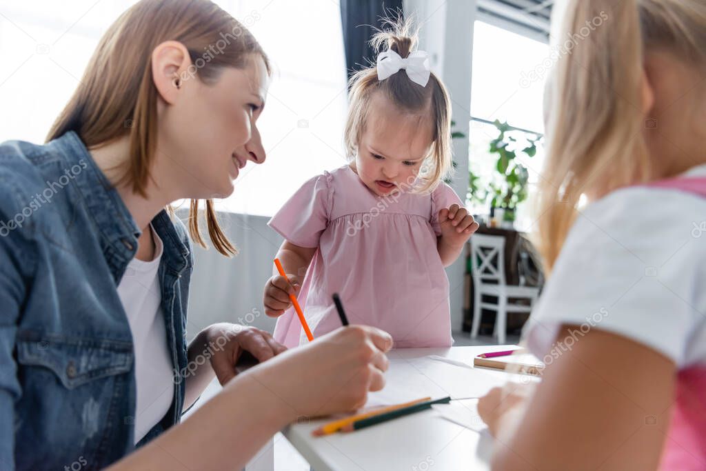 cheerful kindergarten teacher drawing and looking at blurred toddler kid with down syndrome 
