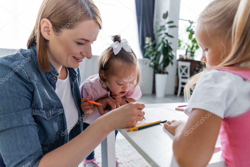 happy kindergarten teacher drawing near toddler kid with down syndrome and preschooler girl