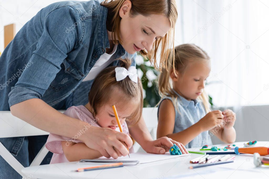 teacher assisting disabled child with down syndrome drawing near blurred child in private kindergarten 