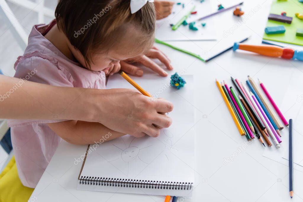 teacher assisting disabled girl with down syndrome drawing in private kindergarten 