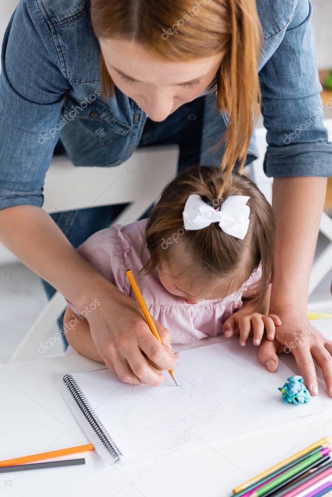 high angle view of teacher assisting disabled girl with down syndrome drawing in private kindergarten 