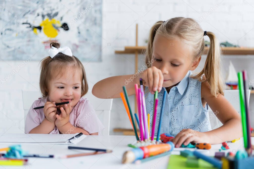 blonde child reaching color pencils near disabled toddler kid with down syndrome in private kindergarten 