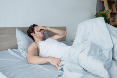 Genç adam yatakta uzanırken gözlerini eliyle kapatıyor.