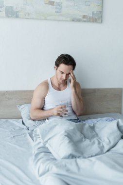 Genç adam bir bardak suyla yatakta oturuyor ve baş ağrısı çekiyor.