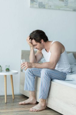 Pijamalı bir adam bir bardak suyla yatakta otururken baş ağrısı çekiyor.