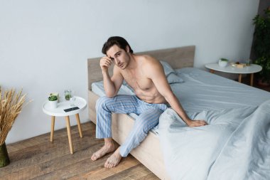 Mavi pijamalı üstsüz adam, sabahları yatakta otururken kendini iyi hissetmiyor.