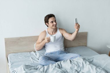 Kahveli gülümseyen adam yatakta cep telefonuyla selfie çekiyor.