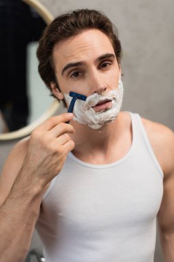 young brunette man looking at camera while shaving in morning clipart