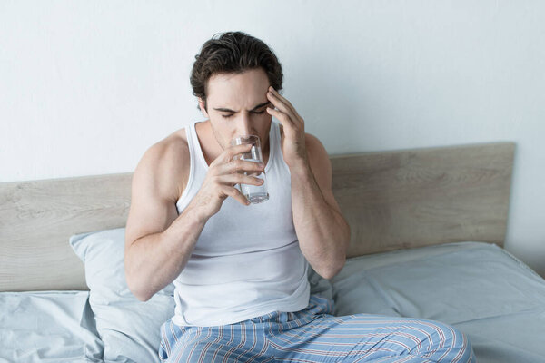 Молодой человек пьет воду с закрытыми глазами, страдая от головной боли