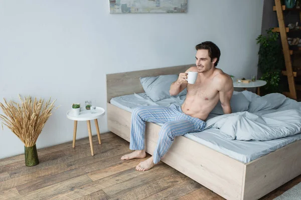 Félmeztelen Férfi Csíkos Pizsama Nadrágban Mosolyog Miközben Kávét Iszik Hálószobában — Stock Fotó