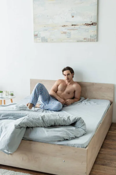 穿着条纹睡衣裤 赤身裸体的男人在床上看着摄像机 — 图库照片