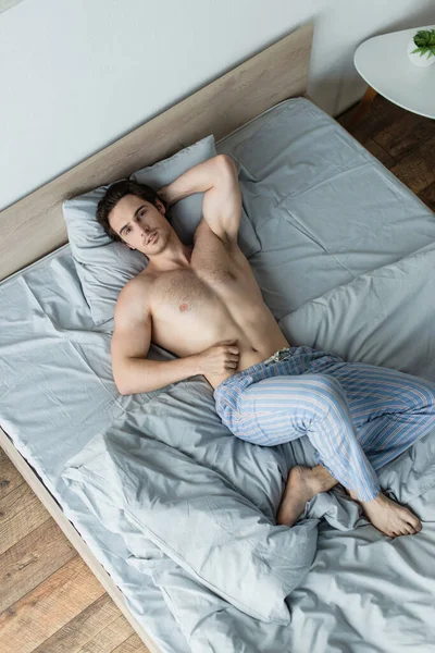 high angle view of shirtless man looking at camera while lying in bed