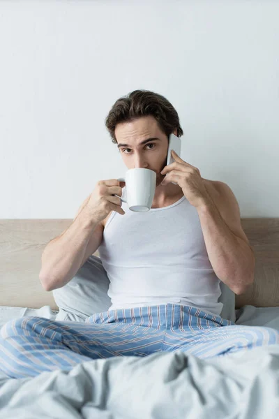 Fiatal Férfi Iszik Kávét Beszél Mobiltelefonnal Miközben Ágyban — Stock Fotó