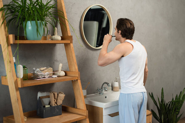 брюнетка мужчина в белом танке топ чистки зубов возле раковины и деревянные стойки в ванной комнате