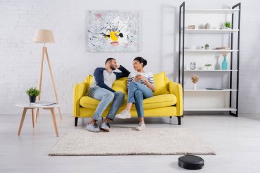 Akıllı telefonları olan ırklar arası genç bir çift robot elektrik süpürgesinin yanında konuşuyor. 