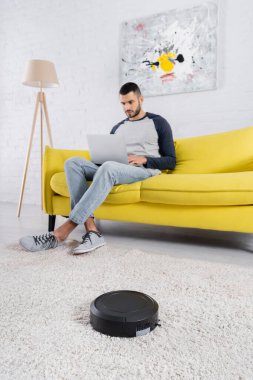 Bulanık adamın yanındaki halının üstünde dizüstü bilgisayar kullanan robot süpürge. 