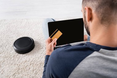 Robotik elektrik süpürgesinin yanında kredi kartı ve laptop tutan bulanık adamın arka görüntüsü 
