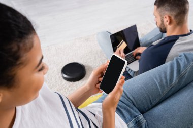 Bulanık Afrikalı Amerikalı kadın erkek arkadaşının yanında akıllı telefon ve evde elektrikli süpürge tutuyor. 