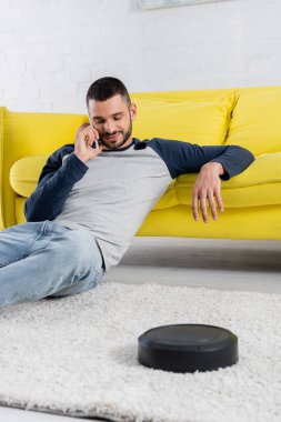 Bulanık robotik elektrik süpürgesinin yanında akıllı telefondan konuşan genç adam. 