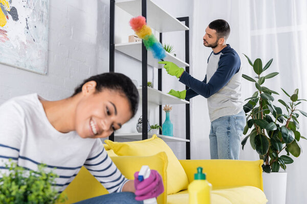 Young man cleaning cupboard with dust brush near blurred african american girlfriend 