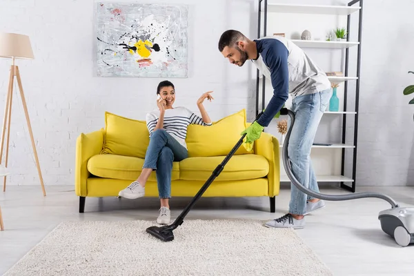 Hombre Aspirando Alfombra Cerca Sonriente Novia Afroamericana Hablando Teléfono Inteligente — Foto de Stock