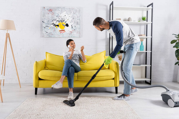 African american woman talking on mobile phone while boyfriend vacuuming carpet 