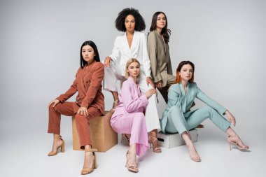 Gri üzerinde otururken şık takım elbiseli ırklar arası modellerin tam uzunluğu