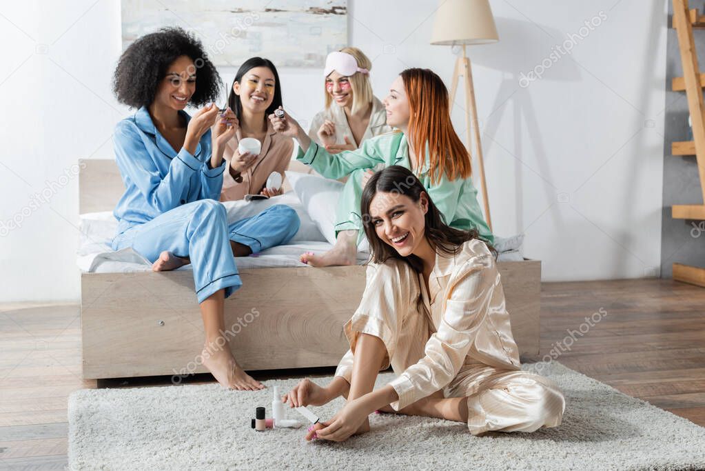 smiling interracial women doing beauty procedures during slumber party in bedroom 