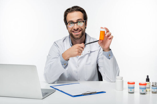 smiling doctor pointing with pen at container with medical cannabis pills isolated on white