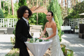 Mosolygó fajok közötti leszbikus pár áll közel csokor és pezsgő esküvő közben szabadban 