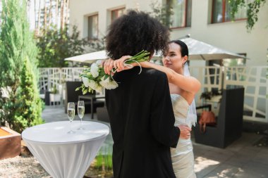 Lesbian woman holding wedding bouquet while hugging african american girlfriend in suit  clipart