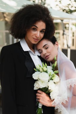 Young lesbian woman in veil holding bouquet near african american girlfriend in suit  clipart