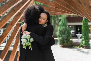 Lesbian woman with wedding bouquet hugging african american girlfriend under arch in park  clipart
