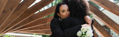 Young lesbian woman holding wedding bouquet and hugging african american girlfriend in park, banner  clipart