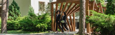 Multiethnic same sex couple with wedding bouquet walking in park, banner  clipart