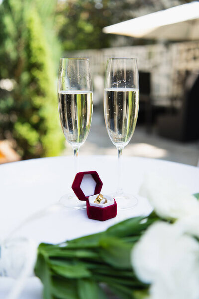 Wedding rings near glasses of champagne and blurred flowers outdoors 
