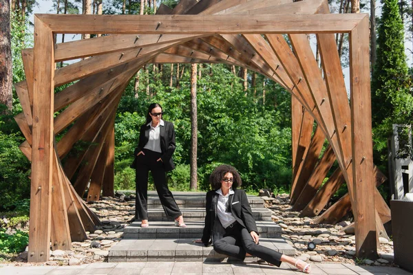 Interracial Lesbian Couple Suits Sunglasses Wooden Arch Park — Stock Photo, Image