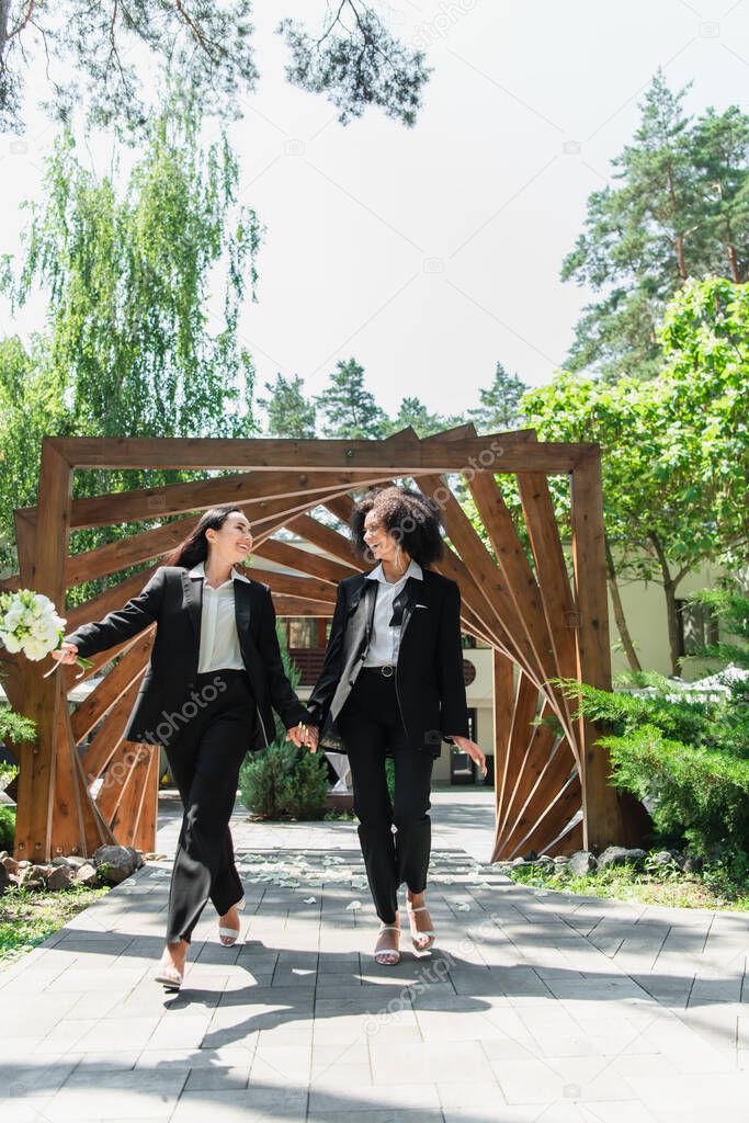 Smiling interracial lesbian couple with wedding bouquet holding hands in park 