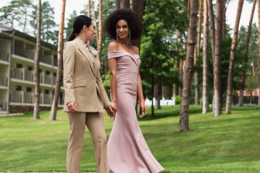 Smiling interracial lesbian couple in dress and suit holding hands while walking in park  clipart