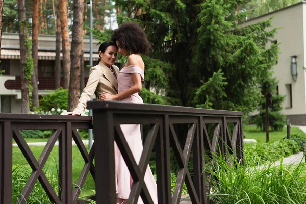 Takım elbiseli, gülümseyen bir kadın. Briçte giyinmiş, Afro-Amerikan kız arkadaşının yanında duruyor. 