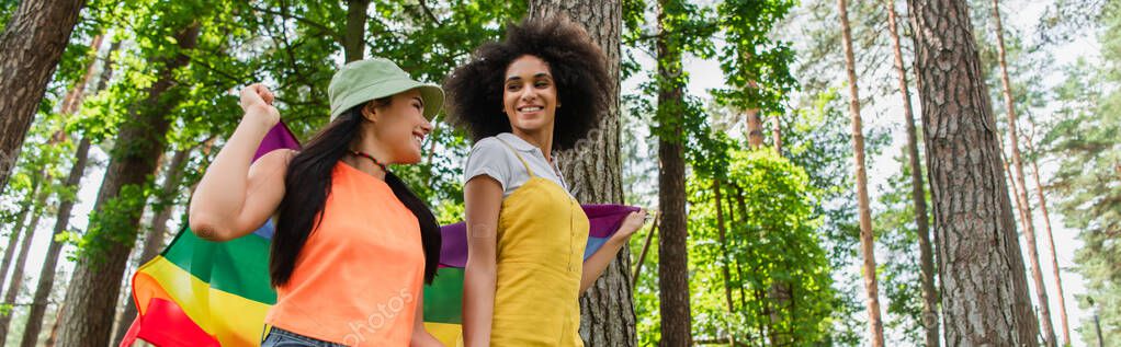 Low angle view of smiling african american woman holding lgbt flag with girlfriend outdoors, banner