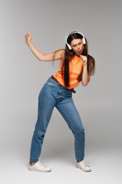 Kot pantolon ve kablosuz kulaklıklı esmer bir kadın. Gri renkte dans ederken müzik dinliyor.
