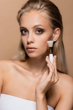 Gri gözlü güzel bir kadın, bej rengi bir yüzün yakınında kozmetik fırça tutuyor.