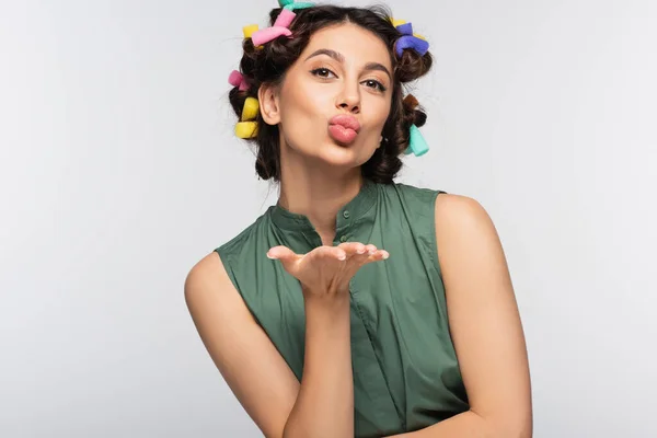 young woman with colorful hair curlers sending air kiss isolated on grey