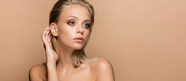 pretty woman with makeup on perfect face touching hair while looking away isolated on beige, banner