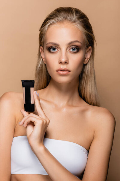 pretty blonde woman with grey eyes showing tube with face foundation isolated on beige