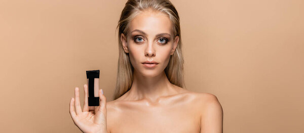 young blonde woman with bare shoulders holding face foundation isolated on beige, banner