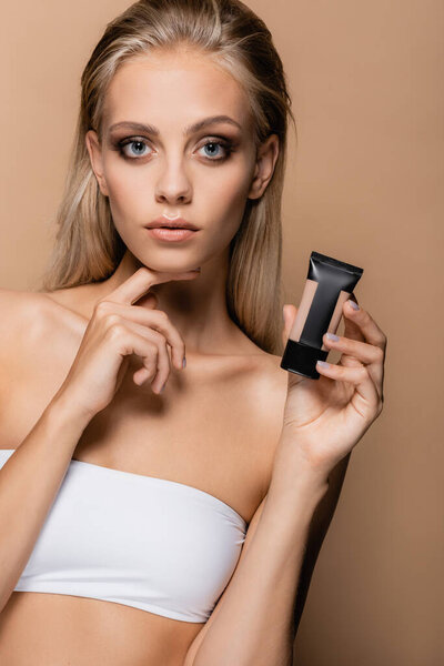 young woman with perfect skin holding face foundation while touching chin isolated on beige